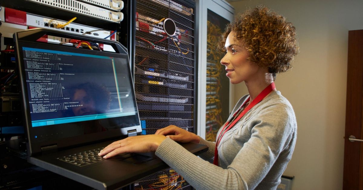 Eine Frau steht neben einem Serverschrank und blickt auf ein Laptop. Auf dessen Display ist Code zu sehen