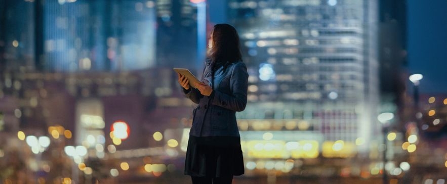 Person mit Tablet vor nächtlicher Stadtkulisse mit verschwommenen Lichtern und Wolkenkratzern im Hintergrund.