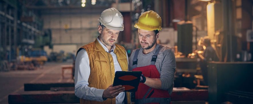 Zwei Bauarbeiter mit Schutzhelmen betrachten ein Tablet in einer Industriehalle, einer trägt gelbe Weste, der andere rote Latzhose.