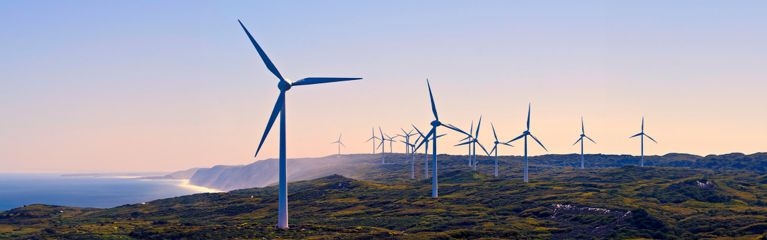 Mehrere Windräder auf grünen Hügeln nahe der Küste bei klarem Himmel, Symbol für nachhaltige Energiegewinnung aus Windkraft