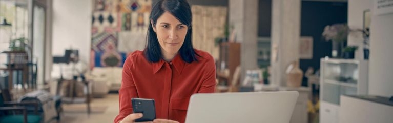Frau mit Smartphone sitzt am Laptop in ihrer Wohnung
