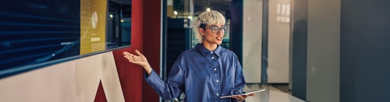 Person mit hellblondem Haar und Brille präsentiert Daten auf großem Bildschirm in modernem Büro, hält Tablet in der Hand