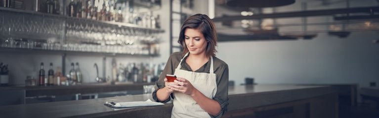 Frau mit Schürze steht mit Smartphone an einer Bar
