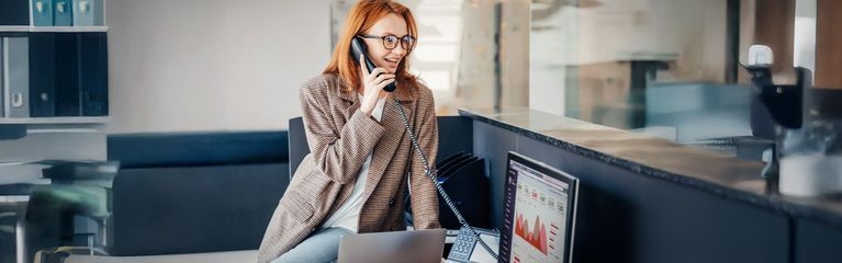 Frau mit Brille arbeitet im Büro in Freizeitkleidung, sitzt auf ihrem Schreibtisch und telefoniert. Moderne, komfortable Büroeinrichtung mit unscharfem Hintergrund. Darstellung einer glücklichen Mitarbeiterin bei der Arbeit.