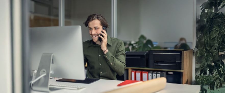 Mann im grünen Hemd telefoniert lächelnd am Arbeitsplatz mit Computer, umgeben von Büropflanzen und Ordnern.