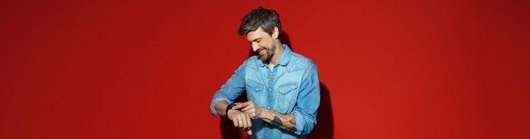 Person in Jeanshemd steht vor rotem Hintergrund und schaut auf eine Smartwatch am Handgelenk