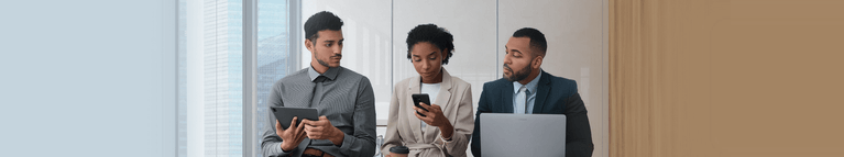 Drei Geschäftsleute in einem hellen Büro mit großen Fenstern, die mit digitalen Geräten arbeiten - Tablet, Smartphone und Laptop.