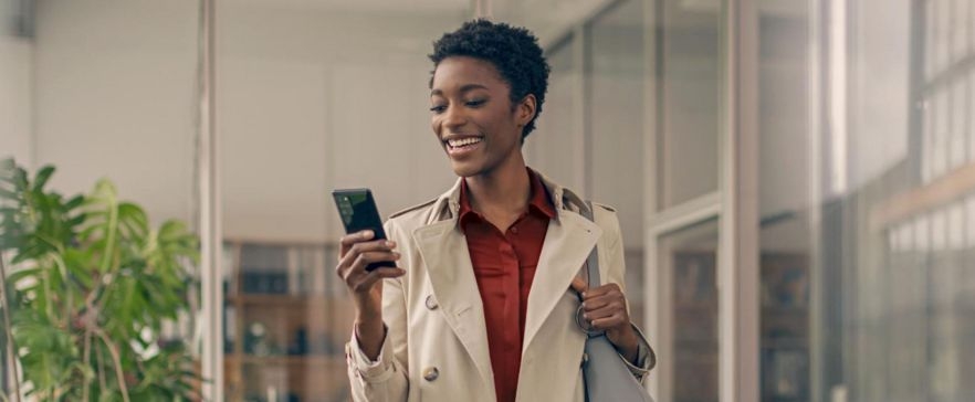 Person in beigem Mantel steht in einem modernen Bürogebäude und hält ein Smartphone in der Hand.