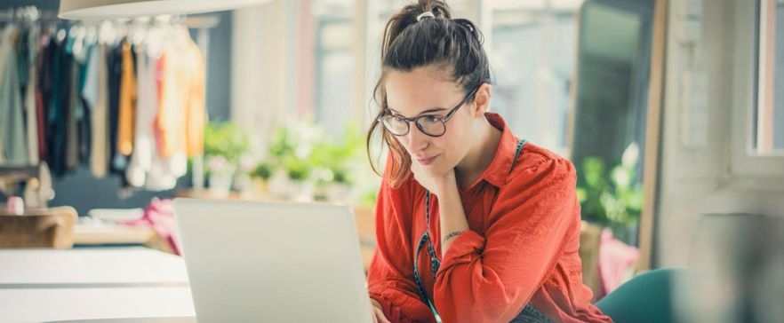 Person sitzt an einem Schreibtisch und arbeitet an einem Laptop in einem hellen Raum mit Kleidung im Hintergrund.