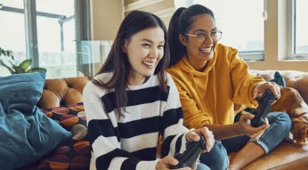 Zwei Freundinnen zocken mit ihren Controllern ein PS4-Spiel.