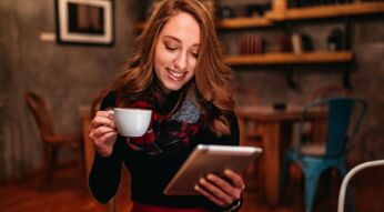 Junge Frau sitzt mit einem Tablet und einer Tasse Kaffee in der Hand auf einem Stuhl