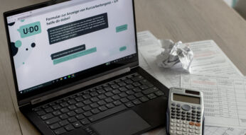 Ein Laptop, auf dessen Bildschirm der Chatbot UDO zur Beantragung von Kurzarbeitergeld zu sehen ist, daneben Unterlagen und ein Taschenrechner.