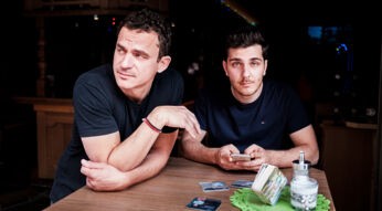 Die Comedy-Autoren Christian Huber und Tarkan Bagci stehen an einem Tisch in einer Kneipe