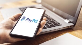 Eine Hand hält ein Smartphone-Display mit dem PayPal-Logo vor einen Laptop.