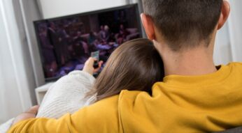 Junge Frau und junger Mann sitzen Arm in Arm auf dem Sofa und gucken Fernsehen