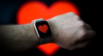 Eine Apple Watch mit rotem Herzsymbol am Handgelenk eines jungen Mannes, for großem Herzsymbol im Hintergrund.