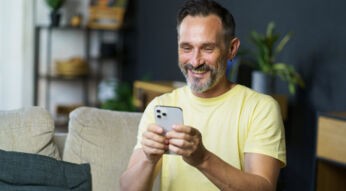 Mann mittleren Alters sitzt lächelnd auf der couch und guckt auf sein iPhone