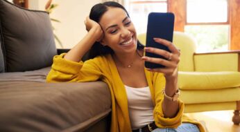 Eine Frau hockt auf dem Boden, hat einen Arm auf der Couch und ein Handy in der Hand