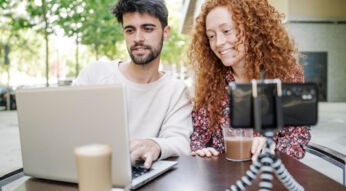 Junger Mann und junge Frau sitzen im Straßencafè am Laptop und filmen sich dabei mit dem Handy.