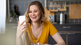 Eine junge Frau sitzt in einer Küche, schaut in ihr Smartphone und lächelt dabei.