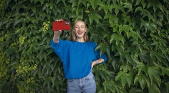 Eine junge Frau steht lächelnd vor einer Efeu-Wand und hält ihr Smartphone in die Kamera