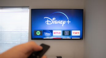 Auf dem Bild ist ein Fernseher zu sehen, auf dem die Benutzeroberfläche mehrerer Streamingdienste angezeigt wird. Im Fokus steht die Disney+ App, die in der Mitte hervorgehoben ist. Eine Hand im Vordergrund hält eine Fernbedienung, bereit zur Auswahl. Das Bild vermittelt eine gemütliche Wohnzimmeratmosphäre und symbolisiert das gemeinsame Streamingerlebnis unter dem Motto Disney+ teilen.