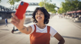 Auf dem Bild in dem Artikel zu den Vodafone CallYa-Tarifen hält eine junge Frau mit lockigen Haaren und orangefarbenem Kleid fröhlich ihr Smartphone in die Höhe, um ein Selfie zu machen. Im Hintergrund ist eine sonnige Fußgängerpromenade mit Bäumen und Cafés zu sehen. Die Szene wirkt lebendig und sommerlich – passend zur frischen Ausstrahlung der Vodafone CallYa-Tarife.