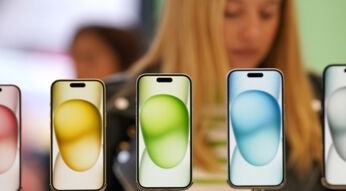 Im Vordergrund mehrere iPhones in der Auslage eines Geschäftes, im Hintergrund guckt eine Frau auf ihr Handy