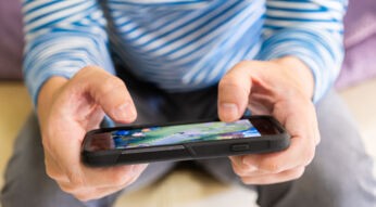 Auf dem Bild zu den besten Handyspielen 2025 hält eine Person mit blau-weiß gestreiftem Shirt ein Smartphone in beiden Händen. Auf dem Bildschirm ist ein farbenfrohes Handyspiel mit grüner Landschaft zu sehen. Die Szene vermittelt eine entspannte Atmosphäre und hebt die Popularität von Mobile Gaming hervor.