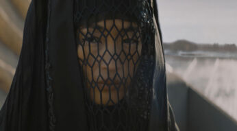 Hier ist dein Alt-Text: "Auf diesem Bild aus Dune Prophecy ist die Figur Tula Harkonnen zu sehen. Sie trägt ein schwarzes, verschleiertes Gewand, das ihr Gesicht bis auf die Augen durch ein netzartiges Muster verdeckt. Ihr Blick wirkt ernst und nachdenklich, während im Hintergrund eine karge, wüstenartige Landschaft zu erahnen ist."