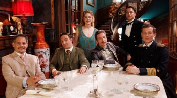 Auf dem Bild aus Dinner For Five sind sechs Personen zu sehen, die in eleganter Kleidung an einem festlich gedeckten Tisch sitzen. Im Hintergrund ist eine aufwendig dekorierte Treppe und ein luxuriös eingerichteter Raum zu erkennen. Die abgebildeten Personen sind Alicia von Rittberg, Kostja Ullmann, Moritz Bleibtreu, Frederick Lau, Jacob Matschenz und Christoph Schechinger.