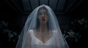 Auf dem Bild aus Something Very Bad is Going to Happen ist Camila Morrone als Rachel Harkin zu sehen. Sie trägt ein weißes Brautkleid mit tiefem Ausschnitt und einen durchsichtigen Schleier, der ihr Gesicht leicht verhüllt. Ihr Blick wirkt ernst und angespannt. Die dunkle, floral geschmückte Umgebung erzeugt eine düstere, bedrohliche Stimmung.