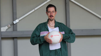 Auf dem Bild zu "Go Präsident Knabe" steht Maximilian "HandOfBlood" als Kunstfigur Präsident Knabe mit einem Trainingsanzug vor einer grauen Strahlträger-Wand. Er schaut etwas deplatziert aus und hält einen Fußball mit der Aufschrift "Vodafone" in den Händen