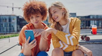 Auf dem Bild in dem Artikel zu Vodafone Young Tarifen sitzen zwei junge Frauen im Freien auf einer Fläche. Sie schauen gemeinsam auf ein Smartphone mit blauer Hülle.