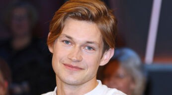 Auf dem Bild ist der deutsche Schauspieler Damian Hardung zu sehen, wie er in die Kamera lächelt. Er hat hellbraune Haare, die leicht zur Seite gestylt sind, und blaue Augen. Sein Gesichtsausdruck wirkt freundlich und entspannt. Der Hintergrund ist unscharf, sodass der Fokus auf seinem Gesicht liegt.