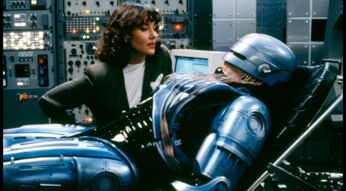 In diesem Szenenbild aus *RoboCop 2* sehen wir die Figur RoboCop in einem High-Tech-Labor auf einer Liege, während er von einer Frau in einem weißen Oberteil und schwarzen Blazer betreut wird. Die Frau schaut aufmerksam auf RoboCop, der in seiner ikonischen metallisch-blauen Rüstung daliegt. Kabel und Schaltkreise sind an seinen Körper angeschlossen, während im Hintergrund eine Vielzahl an Monitoren und Schaltflächen aufleuchtet, was die futuristische und düstere Atmosphäre unterstreicht. Diese Szene zeigt RoboCop in einer ruhenden Position, vermutlich während einer Wartungs- oder Reparatursitzung, und verstärkt die technologische und menschliche Dualität der Figur – ein Thema, das auch in einem modernen RoboCop-Reboot spannend wäre.