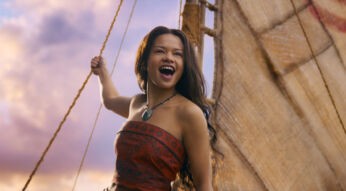 In der Szene aus den Disney Realverfilmungen 2026 sieht man Catherine Laga'aia als Vaiana auf einem traditionellen Segelboot bei Sonnenuntergang. Sie trägt ein rotes, gemustertes Oberteil und eine grüne Muschelkette. Mit offenem Mund und freudigem Blick hält sie sich an einem Seil fest, während das Segel im Hintergrund sichtbar ist. Die Stimmung wirkt abenteuerlich und optimistisch.