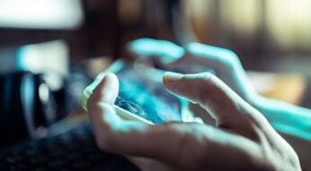 Das Bild in dem Artikel über die besten Koop-Games fürs Handy, zeigt eine Nahaufnahme von Händen, die ein Smartphone im Querformat halten. Der Fokus liegt auf den Fingern und dem Bildschirm, der unscharf, aber lebendig wirkt. Die Atmosphäre ist konzentriert, das Spielgeschehen spiegelt sich leicht auf dem Display wider.