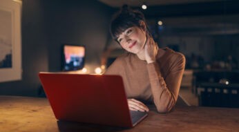Auf dem Bild zum Vodafone Speedtest sitzt eine junge Frau entspannt an einem Holztisch vor einem roten Laptop in einer gemütlich beleuchteten Wohnung. Sie lächelt leicht und blickt konzentriert auf den Bildschirm. Die warme Lichtstimmung und der moderne Wohnraum im Hintergrund vermitteln eine ruhige und angenehme Atmosphäre, passend zu schnellem und komfortablem Internet zuhause.
