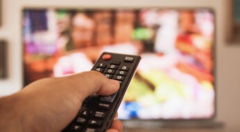 Eine Hand hält eine Fernbedienung, während im Hintergrund ein unscharfer Fernseher mit bunten Inhalten zu sehen ist. Dieses Bild illustriert das Streaming-Erlebnis, passend zum Thema Crunchyroll Kosten 2025.