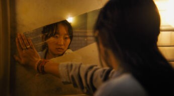 Im Bild zum Until Dawn-Film ist Megan, gespielt von Ji-young Joo, zu sehen. Sie blickt ernst in einen schmutzigen Spiegel, während ihre Hand gegen die spiegelnde Oberfläche drückt. Die Szene ist in warmes, gedämpftes Licht getaucht, was eine nachdenkliche und emotionale Stimmung erzeugt.