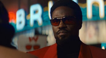 In dieser Szene aus der Wonder Man-Serie ist Yahya Abdul-Mateen II als Simon Williams zu sehen. Er trägt ein auffälliges rotes Sakko, dunkle Sonnenbrille und blickt ernst in die Ferne.