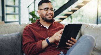 Junger Mann sitzt auf der Couch und arbeitet an seinem Tablet.