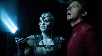 Auf dem Bild aus Star Trek Beyond, einem der Filme von Simon Pegg ist der Schauspieler als Montgomery "Scotty" Scott in einer düsteren Szene zu sehen. Neben ihm steht Jaylah, gespielt von Sofia Boutella, mit weißer Haut und schwarzen Gesichtsmustern. Sie hält eine futuristische Taschenlampe, die ein starkes blaues Licht ausstrahlt. Beide blicken gespannt nach oben, als ob sie etwas Unbekanntes entdeckt hätten. Die dunkle Umgebung und die Beleuchtung erzeugen eine geheimnisvolle und angespannt wirkende Atmosphäre.