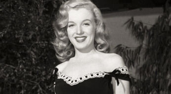 Auf dem Bild, das symbolisch für Dokus über Frauen zum Weltfrauentag verwendet wird, ist Marilyn Monroe in einer eleganten Pose zu sehen. Sie trägt ein schwarzes, schulterfreies Kleid mit Spitzenverzierung und Schleifen. Ihr blondes Haar fällt in sanften Wellen, und sie strahlt mit einem freundlichen Lächeln. Der Hintergrund ist unscharf und zeigt Pflanzen, was das Bild natürlich wirken lässt.