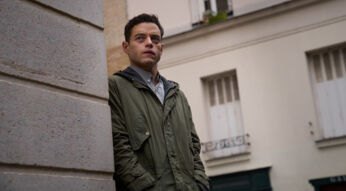 Auf dem Bild aus The Amateur ist Charles Heller zu sehen, gespielt von Rami Malek. Er steht in einer Straßenszene an einer Häuserwand und blickt mit ernstem, nachdenklichem Gesichtsausdruck in die Ferne. Sein olivgrüner Parka und die gedeckten Farben der Umgebung verleihen der Szene eine gespannte, geheimnisvolle Stimmung. Das Bild vermittelt einen stillen Moment voller innerer Anspannung und Ungewissheit.