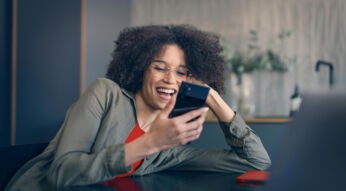 Auf dem Bild zur Vodafone Kombi stützt sich eine lachende Frau mit lockigem Haar und Brille entspannt auf einem Tisch ab. Sie hält ein schwarzes Smartphone vor sich und blickt begeistert auf den Bildschirm. Ihre lässige Kleidung, das moderne Wohnzimmer und die warme, natürliche Beleuchtung vermitteln eine fröhliche, ungezwungene Stimmung und digitalen Komfort im Alltag.