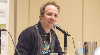 Auf dem Bild in dem Artikel zu dem Big Bang Theory-Spin-off „Stuart Fails to Save the Universe“ ist Kevin Sussman zu sehen, der Stuart in der Serie spielt. Er sitzt lächelnd bei einem Panel hinter einem Mikrofon, trägt ein dunkelblaues Hemd und eine Wettergrafik auf dem T-Shirt. Die Atmosphäre ist locker und freundlich.
