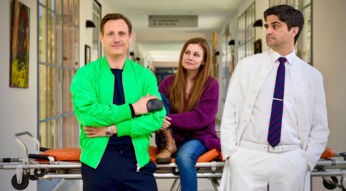 Auf dem Bild zur Serie Dr. Nice Staffel 4 stehen drei Hauptfiguren in einem Krankenhausflur. Links steht Dr. Nice, gespielt von Patrick Kalupa, in einer auffälligen grünen Jacke und lächelt selbstbewusst. In der Mitte sitzt Charlie Winkler, dargestellt von Josefine Preuß, locker auf einer Trage und blickt freundlich. Rechts lehnt Dr. Florian Schmidtke, gespielt von Maximilian Grill, in einem weißen Arztkittel mit verschränkten Armen und skeptischem Blick.
