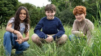 Auf dem Bild zur neuen Harry Potter-Serie sind drei Kinder in einer Wiesenlandschaft zu sehen. In der Mitte sitzt ein Junge mit dunkelblauem Pullover, links von ihm kniet ein Mädchen mit langen dunklen Haaren und weißem T-Shirt, rechts sitzt ein rothaariger Junge in einem beigefarbenen Pullover.